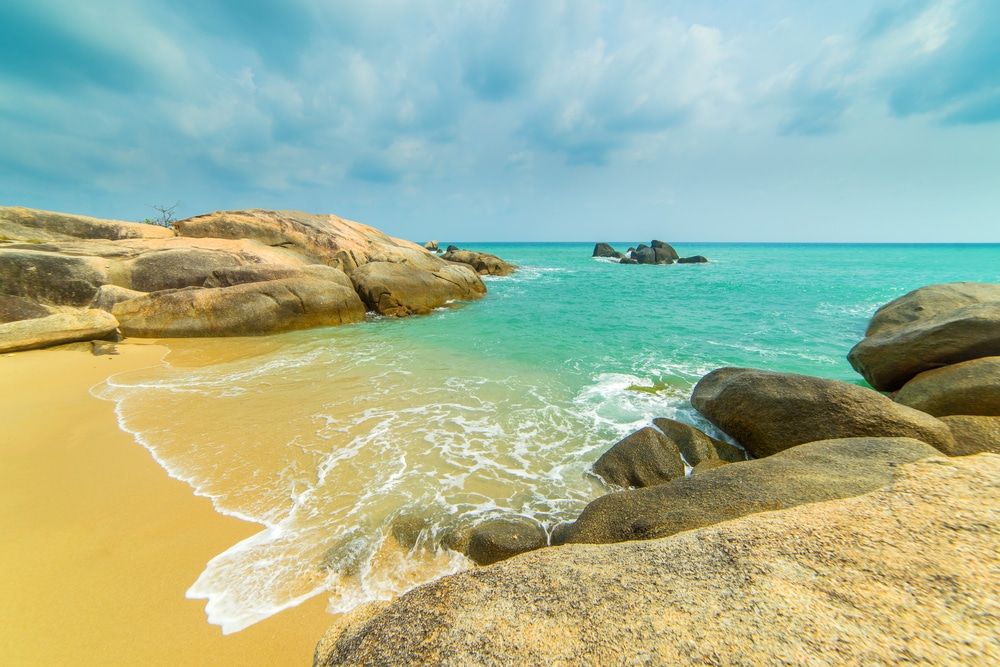 แนวชายฝั่งหินที่มีหาดทรายสีทอง คลื่นสีฟ้าคราม และก้อนหินขนาดใหญ่ที่กระจัดกระจายอยู่ใต้ท้องฟ้าที่มีเมฆบางส่วน เป็นสถานที่อันงดงามสำหรับทุกคนที่กำลังมองห เที่ยวเกาะสมุย
