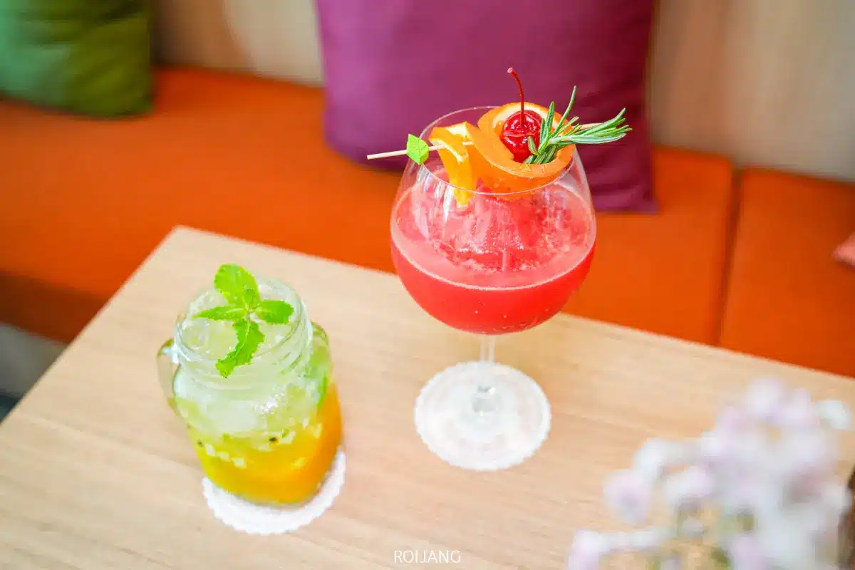 ค็อกเทล Spice House สีสันสดใสสองแก้วบนโต๊ะไม้ แก้วหนึ่งเป็นเครื่องดื่มสีเหลืองเขียวผสมสะระแหน่ในขวด และเครื่องดื่มสีแดงผสมผลไม้ในแก้วทรงก้าน.