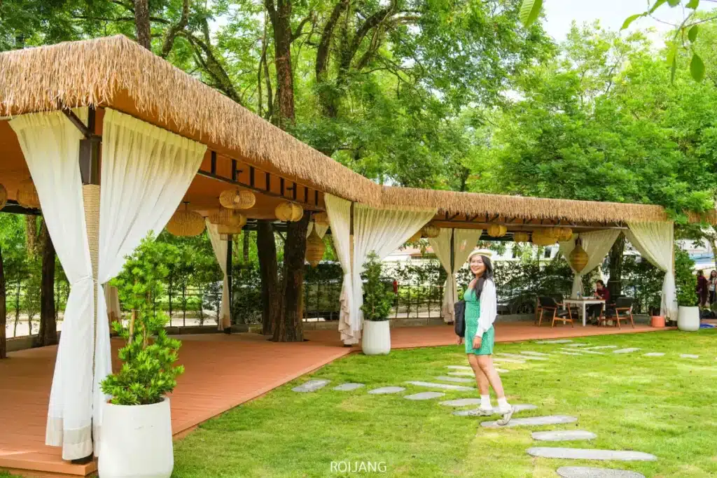 ผู้หญิงในชุดสีเขียวยืนอยู่บนทางเดินหินหน้า Spice House ศาลาแบบเปิดโล่งที่มีหลังคาฟาง ม่านสีขาว และต้นไม้เขียวขจีที่รายล้อมอยู่โดยรอบ.