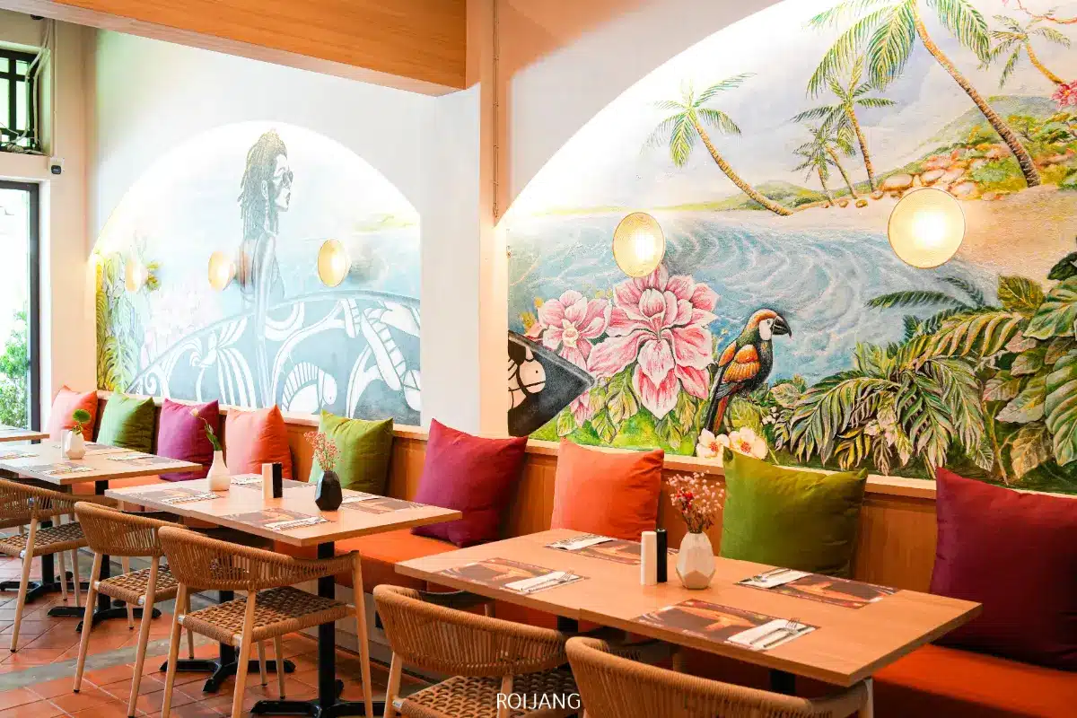 ภายในร้านอาหารสีสันสดใสที่ร้าน Spice House ที่มีโต๊ะไม้ เบาะหลากสี เก้าอี้หวาย และภาพจิตรกรรมฝาผนังขนาดใหญ่ที่มีดอกไม้ นกแก้ว มหาสมุทร และผู้หญิง.