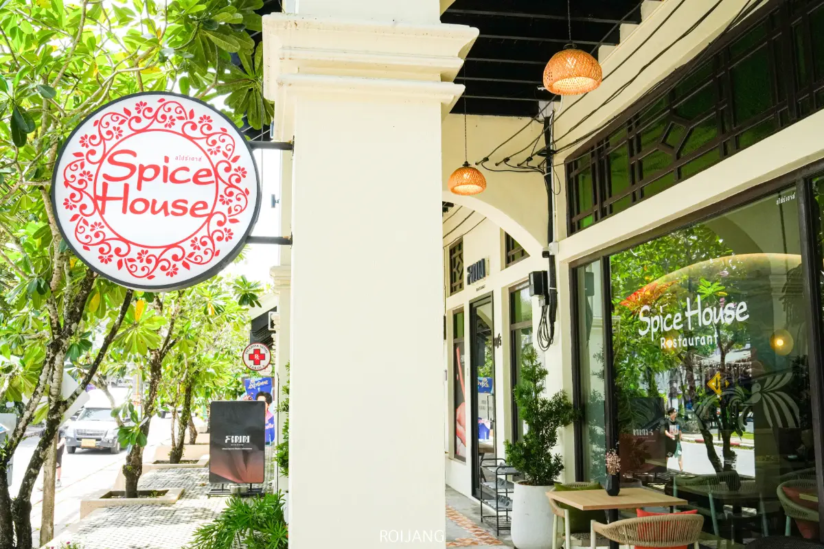 มุมมองถนนของร้านอาหาร Spice House พร้อมป้ายแขวนประดับตกแต่ง หน้าต่างบานใหญ่ และกระถางต้นไม้ ต้นไม้เรียงรายตามทางเท้าในวันที่อากาศแจ่มใส เน้นย้ำถึงบรรยากาศอันน่าดึงดูดของร้าน Spice House.