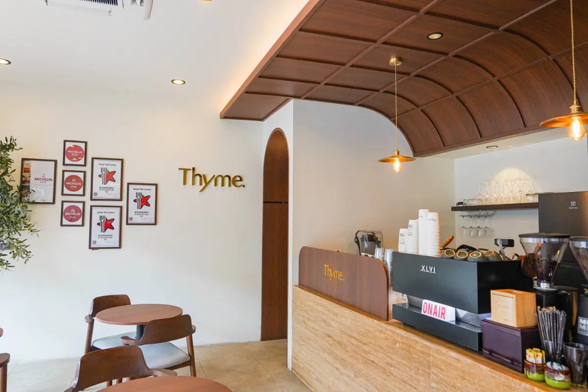 คาเฟ่สไตล์โมเดิร์นที่มีโต๊ะและเก้าอี้ไม้ เคาน์เตอร์กาแฟพร้อมอุปกรณ์ และป้าย "THYME" ที่โดดเด่นบนผนัง รูปภาพใส่กรอบแขวนอยู่ข้างป้าย THYME ช่วยเพิ่มเสน่ห์ให้กับร้าน.