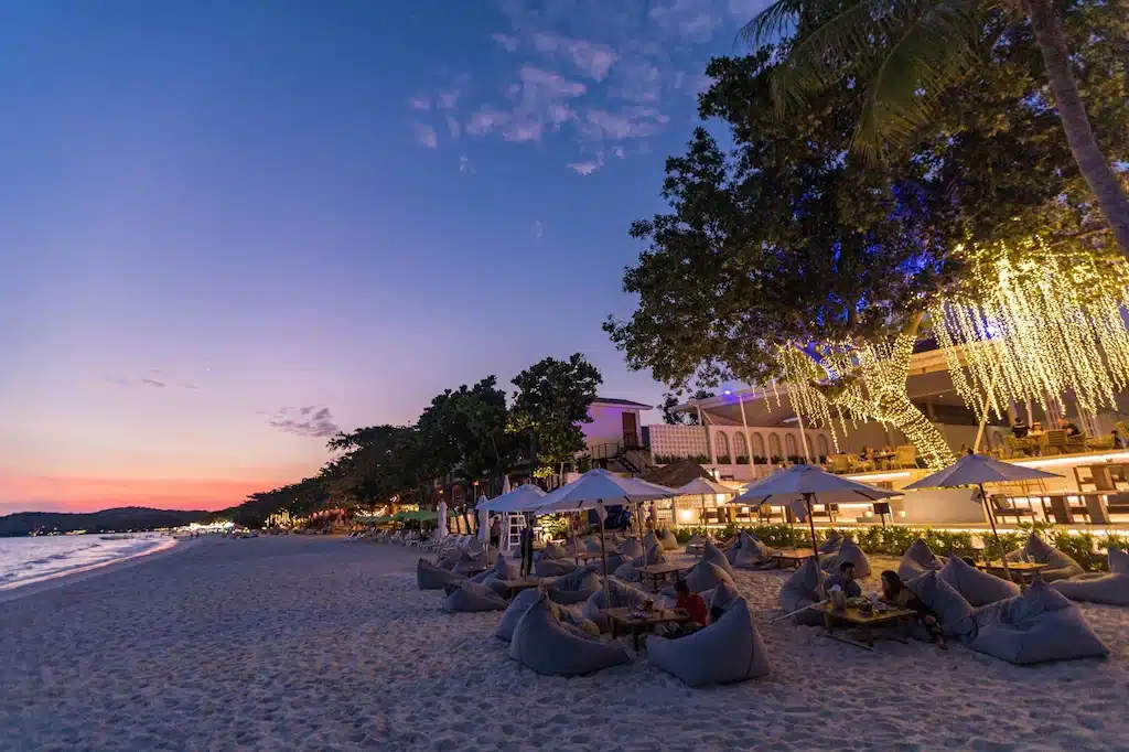 ร้านอาหารริมชายหาดที่มีที่นั่งกลางแจ้งบนเก้าอี้บีนแบ็ก ส่องสว่างด้วยไฟประดับ ขณะพระอาทิตย์ตกดินพร้อมกับผู้คนรับประทานอาหารและทะเลอันสงบอยู่เบื้องหลัง จุดที่ห้ามพลาดสำหรับผู้ที่กำลังมองหา ที่เที่ยวเกาะเสม็ด