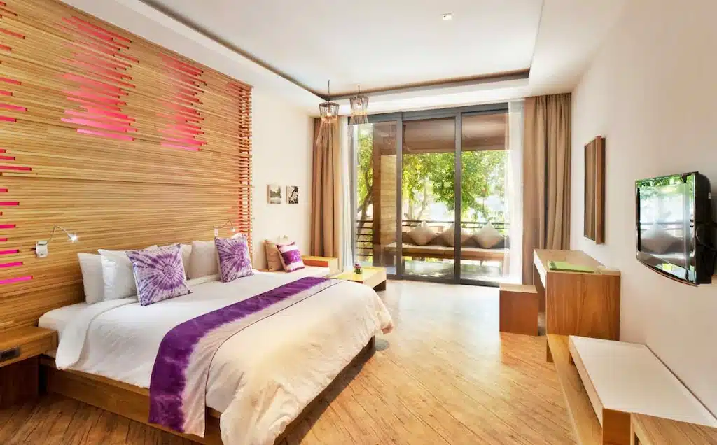 ห้องพักในโรงแรมทันสมัยพร้อมเตียงขนาดใหญ่ ตกแต่งด้วยสีม่วง พื้นไม้ และหน้าต่างสูงจากพื้นจรดเพดานที่ เที่ยวเกาะเสม็ด มองเห็นวิวต้นไม้และลานบ้าน เหมาะสำหรับการพักผ่อนหลังจากเที่ยวชมเกาะเสม็ด.