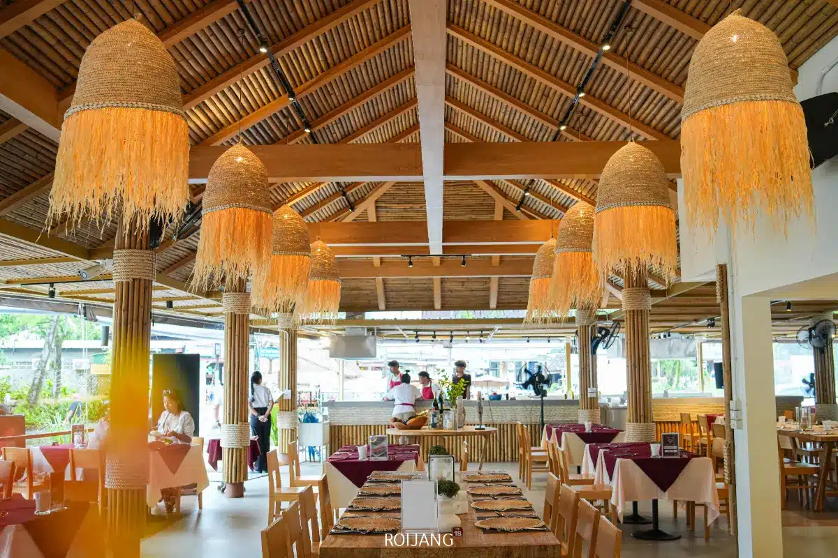 ร้านอาหารรอยเลย์กว้างขวาง ตกแต่งด้วยไม้ โคมไฟฟางแขวนขนาดใหญ่ โต๊ะรับประทานอาหาร และพนักงานที่ยืนอยู่ใกล้เคาน์เตอร์ด้านหลัง.
