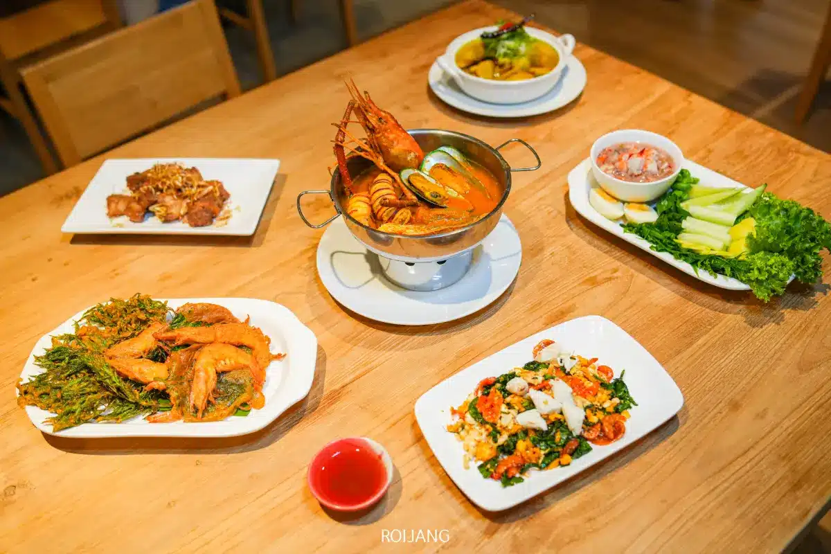 โต๊ะไม้ที่ร้าน Roilay เซ็ตอาหารไทย 5 อย่าง ได้แก่ ต้มซีฟู้ด ปีกไก่ทอด ผัดผัก แกงเหลือง และผักสดพร้อมน้ำจิ้ม.