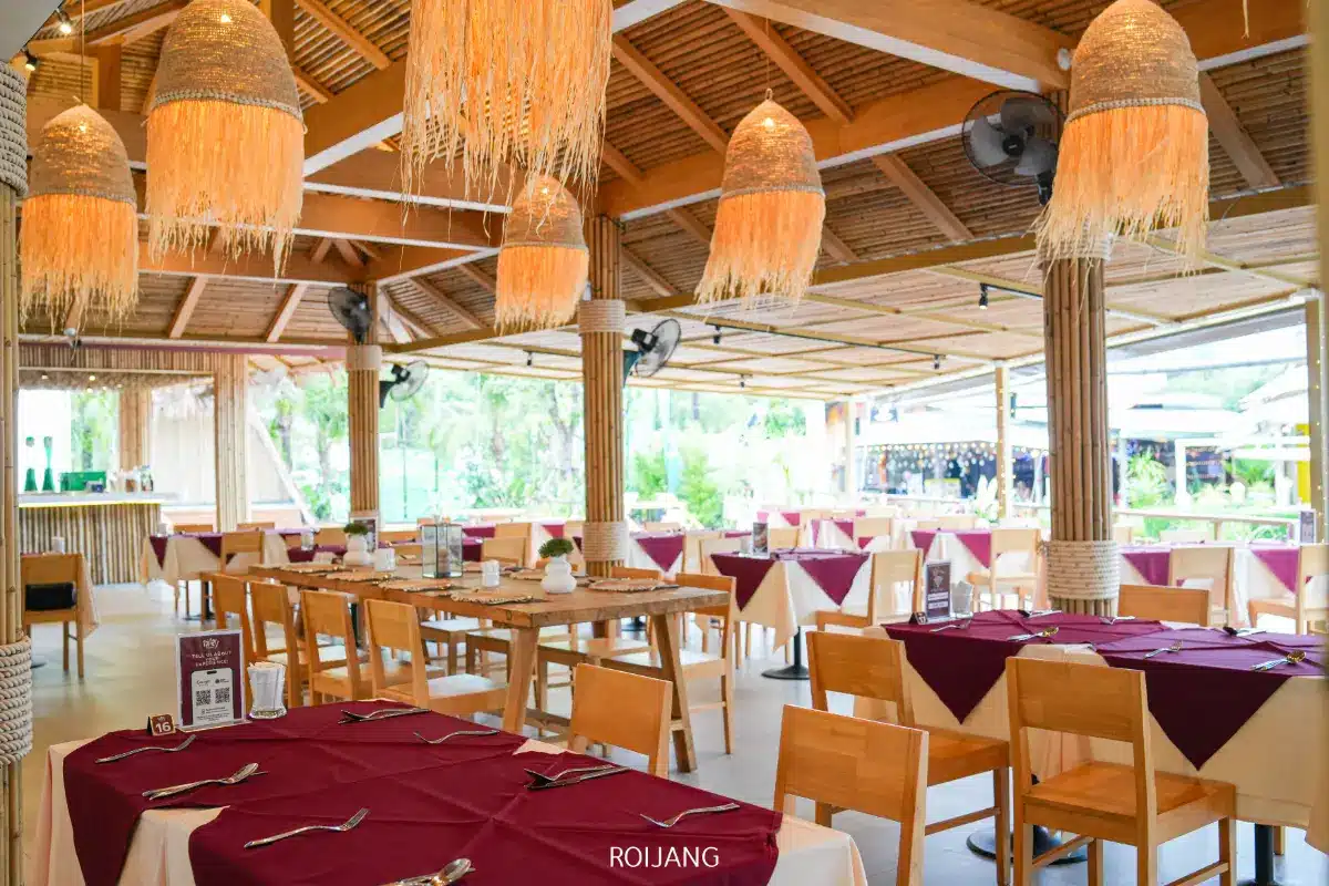 ร้านอาหาร Roilay ตกแต่งภายในกว้างขวาง มีเฟอร์นิเจอร์ไม้ ผ้าปูโต๊ะสีแดงเลือดหมูและสีขาว และโคมไฟทอที่แขวนอยู่ใต้เพดานไม้ไผ่.