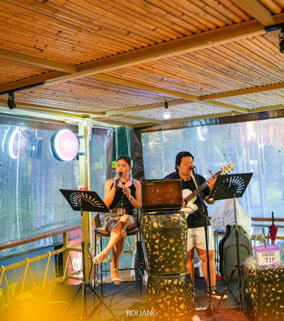 ผู้หญิงคนหนึ่งร้องเพลงเข้าไมโครโฟนขณะนั่งอยู่ และผู้ชายคนหนึ่งเล่นกีตาร์และร้องเพลง ทั้งสองแสดงบนเวทีเล็กๆ ในพื้นที่ Roilay กลางแจ้งที่มีหลังคา.