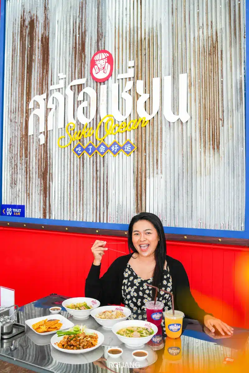 ผู้หญิงคนหนึ่งนั่งอยู่ที่โต๊ะที่มีจานและเครื่องดื่มหลากหลายหน้าป้ายโลหะลูกฟูกที่เขียนว่า “สุกี้โอเชี่ยน”.