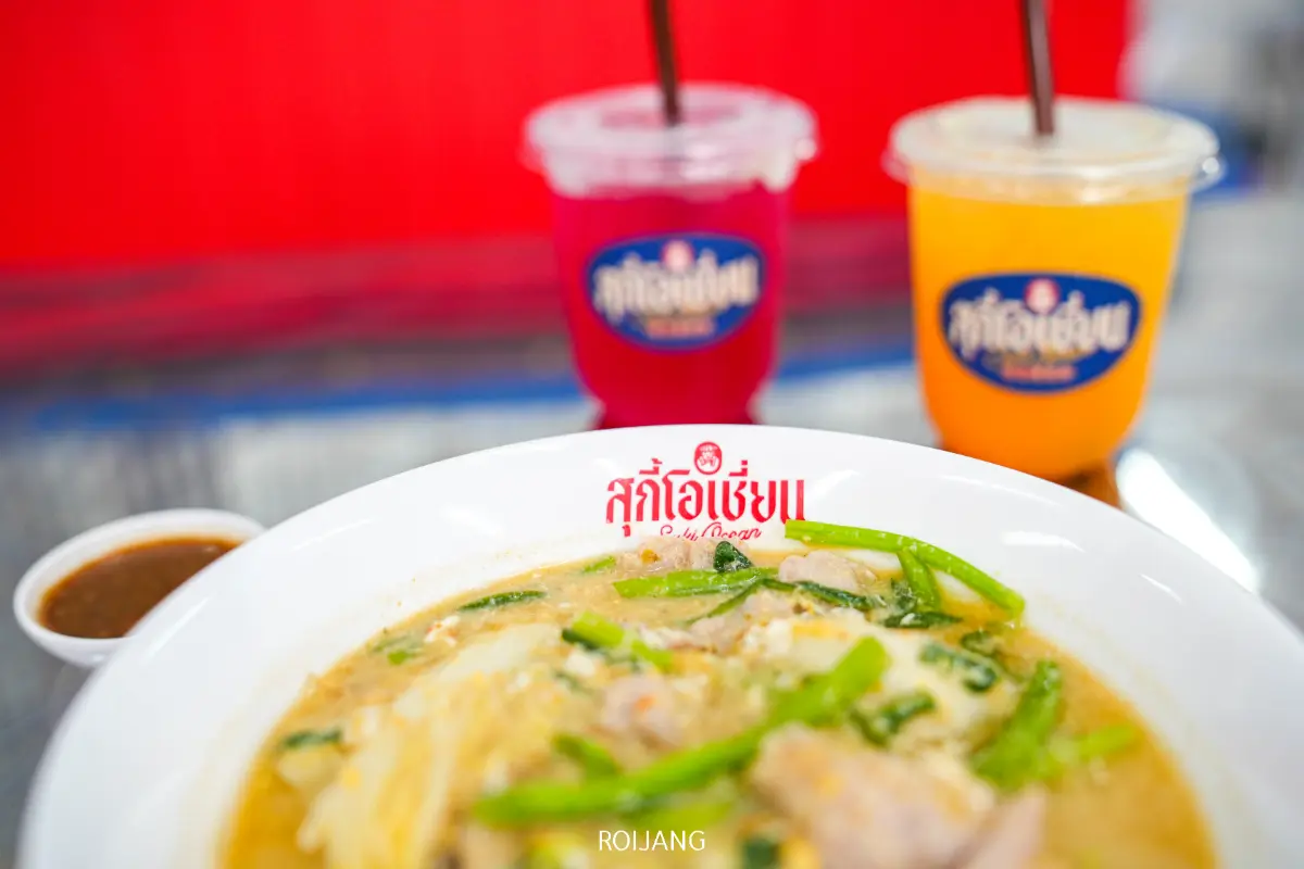 ก๋วยเตี๋ยวสุกี้โอเชี่ยนชามขาวใส่ผักและเนื้อสัตว์เป็นจุดสนใจ พร้อมด้วยเครื่องดื่มสีสันสดใสสองแก้วและซอสจานเล็ก ๆ อยู่เบื้องหลัง.
