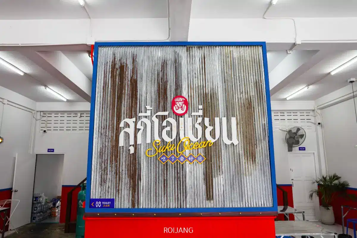ป้ายโลหะลูกฟูกขนาดใหญ่พร้อมข้อความภาษาไทยและภาษาอังกฤษ เช่น “สุกี้โอเชี่ยน” และ “สุกี้โอเชี่ยน” วางอยู่ภายในห้องที่มีแสงสว่างสดใสด้วยผนังสีแดงและสีน้ำเงิน.
