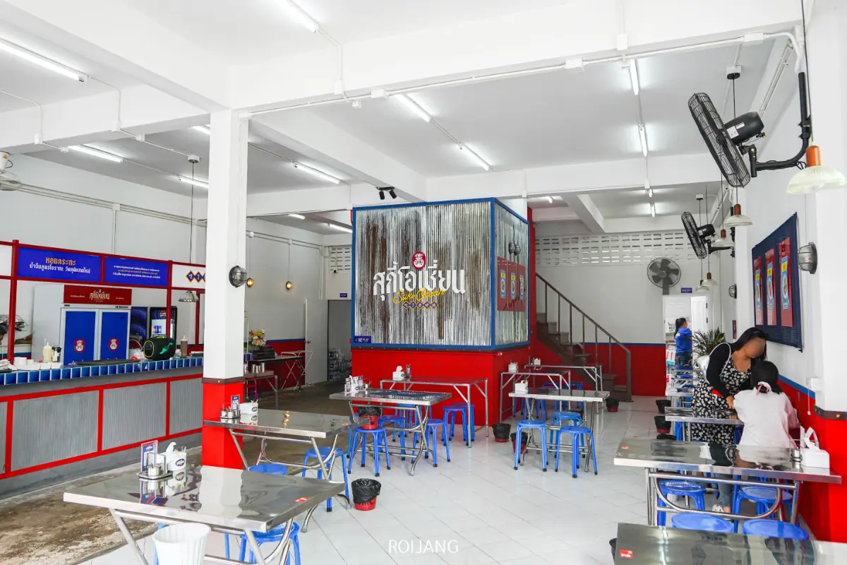 ร้านอาหารสุกี้โอเชี่ยนเป็นร้านอาหารที่สะอาดและมีแสงสว่างเพียงพอ โดยมีโต๊ะและเก้าอี้โลหะสีน้ำเงิน ผนังสีแดงและสีขาว และมีคนนั่งอยู่ไม่กี่คนบริเวณเคาน์เตอร์ทางด้านขวา.