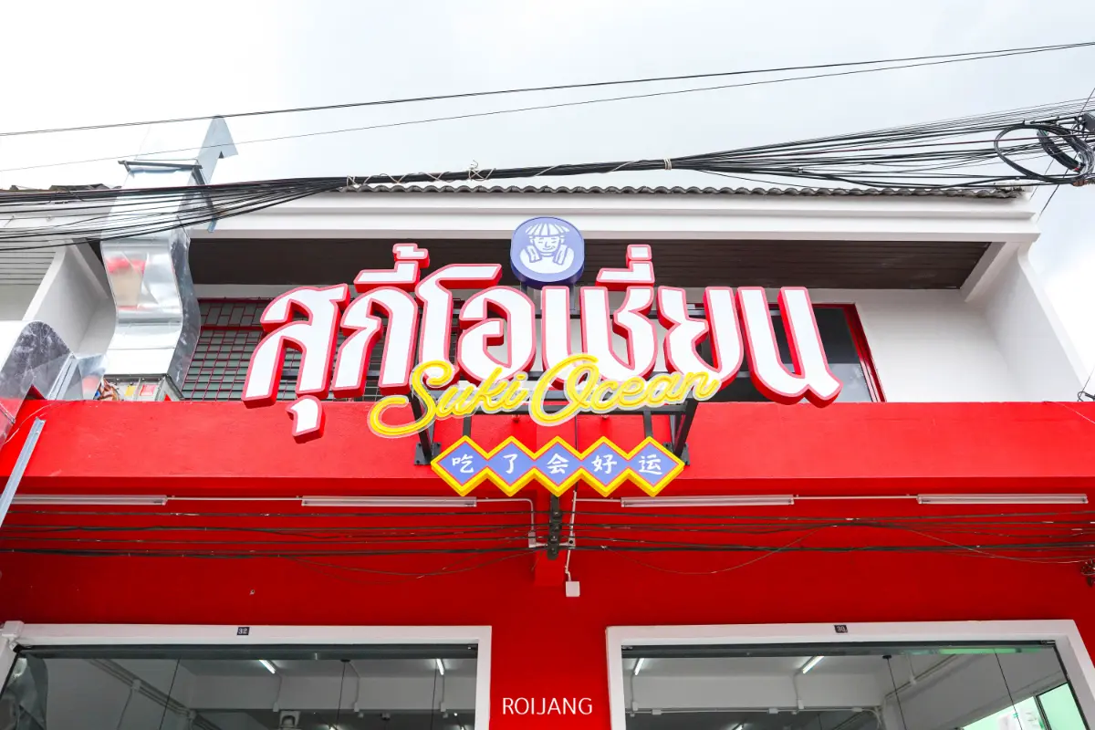 หน้าร้านสีแดงและสีขาวพร้อมป้ายขนาดใหญ่ระบุว่า "สุกี้โอเชี่ยน" เป็นภาษาอังกฤษ ภาษาไทย และอักษรจีนเพิ่มเติมด้านล่าง.