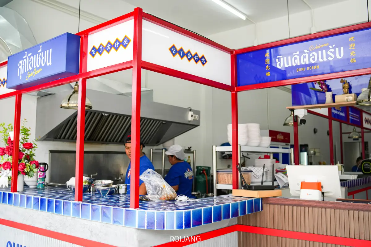 คนสองคนทำงานอยู่ด้านหลังเคาน์เตอร์กระเบื้องสีฟ้าในครัวร้านอาหารที่มีแสงสว่างสดใส มีป้ายภาษาไทยและชื่อ สุกี้โอเชี่ยน แสดงอยู่เหนือบริเวณทำอาหาร.