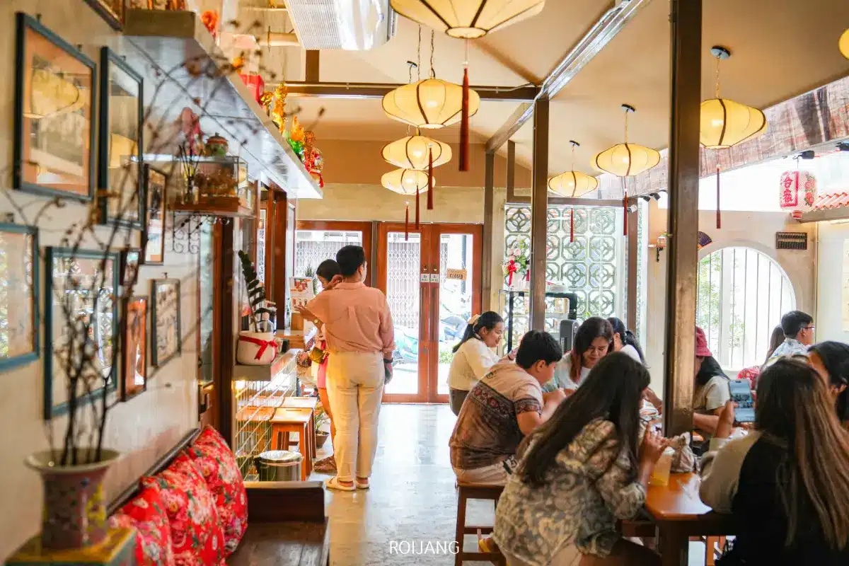 ร้านอาหารโรงโกปี้ที่มีแสงสว่างสดใส ผู้รับประทานอาหารนั่งที่โต๊ะไม้ ผู้คนกำลังรับประทานอาหารและพูดคุยกัน และพนักงาน 2 คนยืนอยู่ใกล้ทางเข้า.