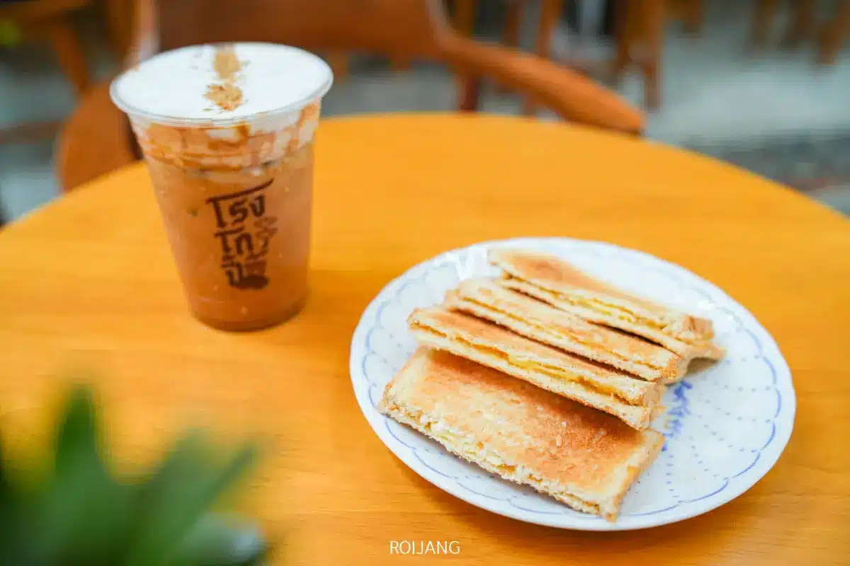 กาแฟเย็นในแก้วพลาสติกและจานแซนวิชปิ้งวางอยู่บนโต๊ะไม้กลมที่โรงโกปี้.