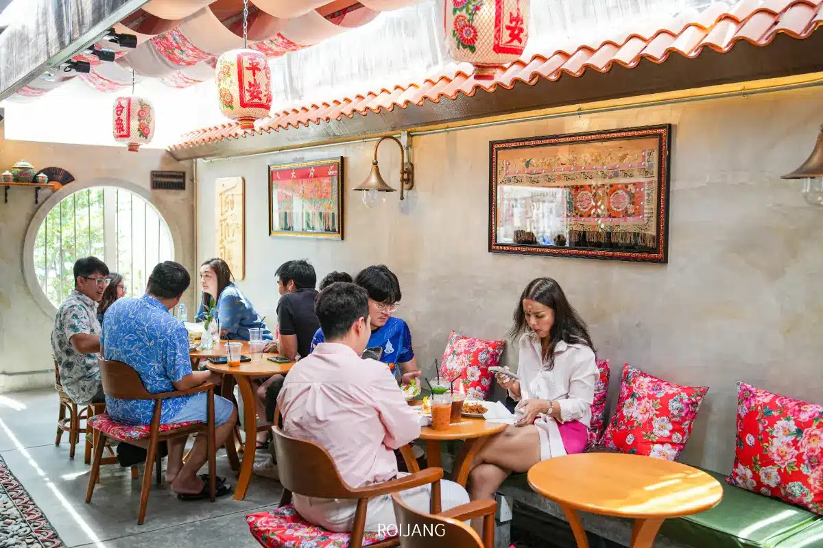 กลุ่มคนนั่งอยู่ที่โต๊ะเล็กๆ ในร้านกาแฟร้านโกปี้ที่มีแสงสว่างสดใส ตกแต่งแบบเอเชียน เช่น โคมไฟสีแดงและเบาะรองนั่งที่มีลวดลายต่างๆ.