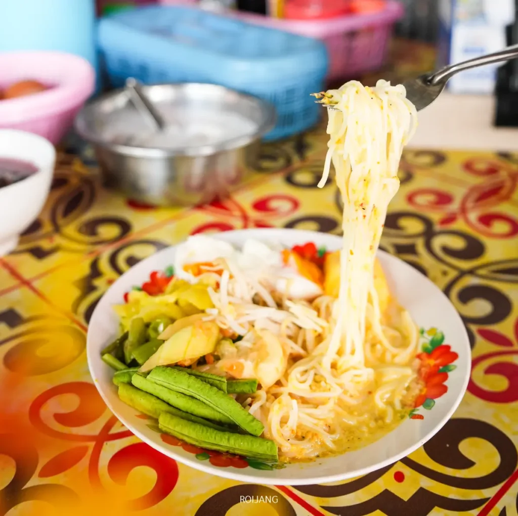 จานก๋วยเตี๋ยวราดซอสแกงเหลืองพร้อมผัก มีคนกำลังยกขนมจีนอี๋น้อง ก๋วยเตี๋ยว โดยใช้ส้อม มีโต๊ะสีสันสดใสและของใช้ในครัวอยู่เบื้องหลัง.