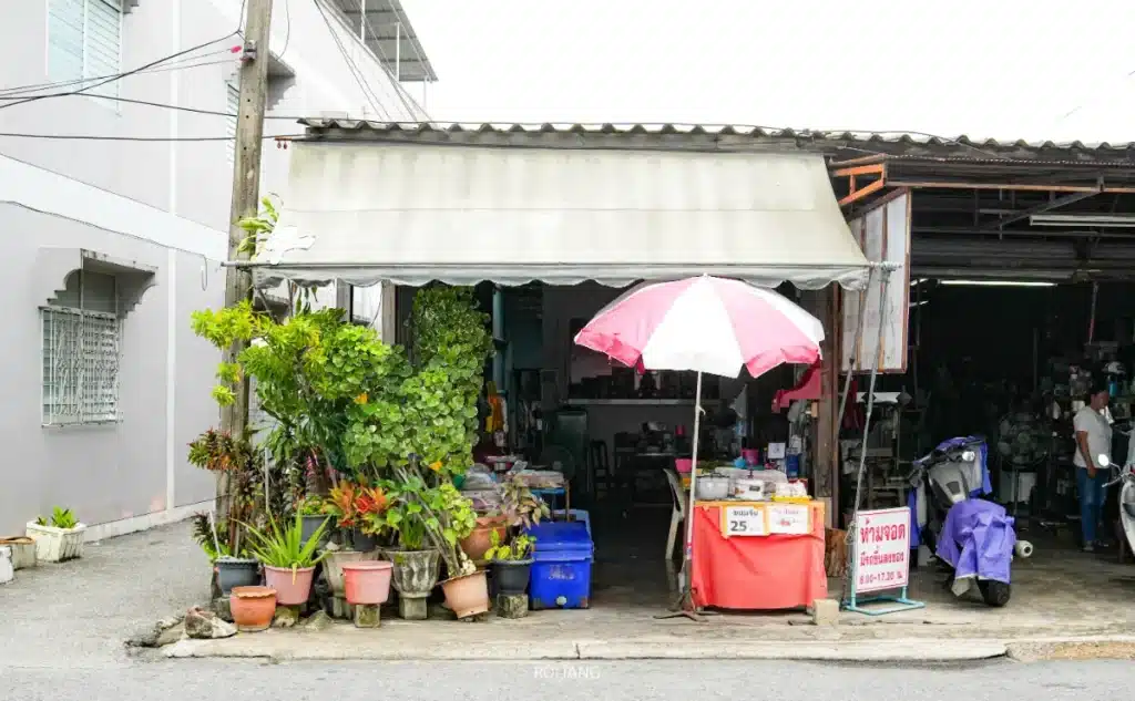 ร้านเล็กๆ แบบเปิดโล่งที่มีกระถางต้นไม้อยู่ด้านหน้า ร่มสีชมพูขาว โต๊ะด้านใน และป้ายโฆษณาขนมจีนอี๋น้อง และอาหาร 25 หน่วยสกุลเงิน.