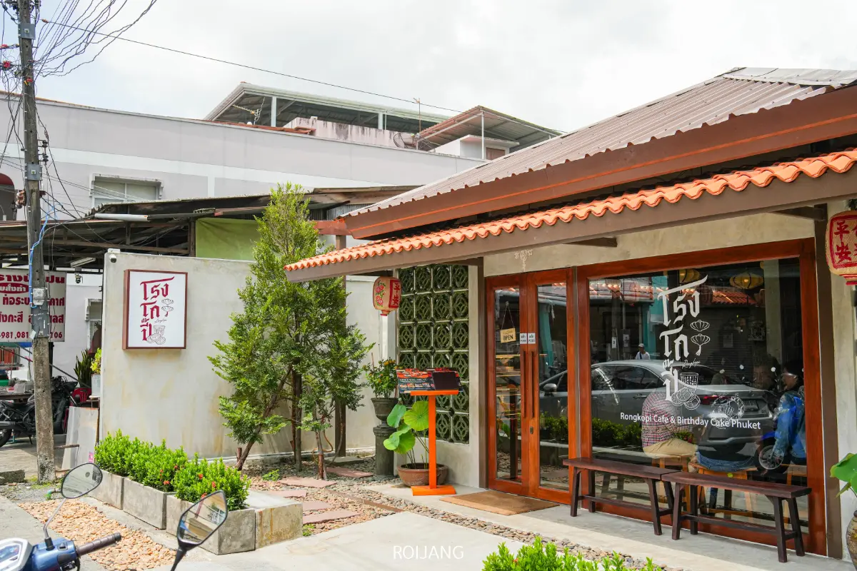คาเฟ่ทันสมัยที่มีหน้าต่างกระจกบานใหญ่ ม้านั่งไม้ และกระถางต้นไม้ PALME ที่ทางเข้า ตั้งอยู่ที่มุมถนนซึ่งมีอาคารและรถจักรยานยนต์จอดอยู่ใกล้เคียง.