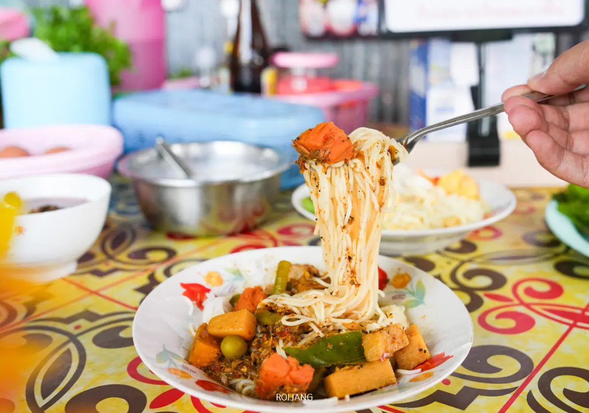 ส้อมยกเส้นก๋วยเตี๋ยวผักและเต้าหู้จากจานสีสันสดใสบนโต๊ะที่มีลวดลาย รายล้อมไปด้วยอุปกรณ์เครื่องครัว เป็นอาหารขนมจีนอี๋น้องแสนอร่อยที่พร้อมเพลิดเพลิน.