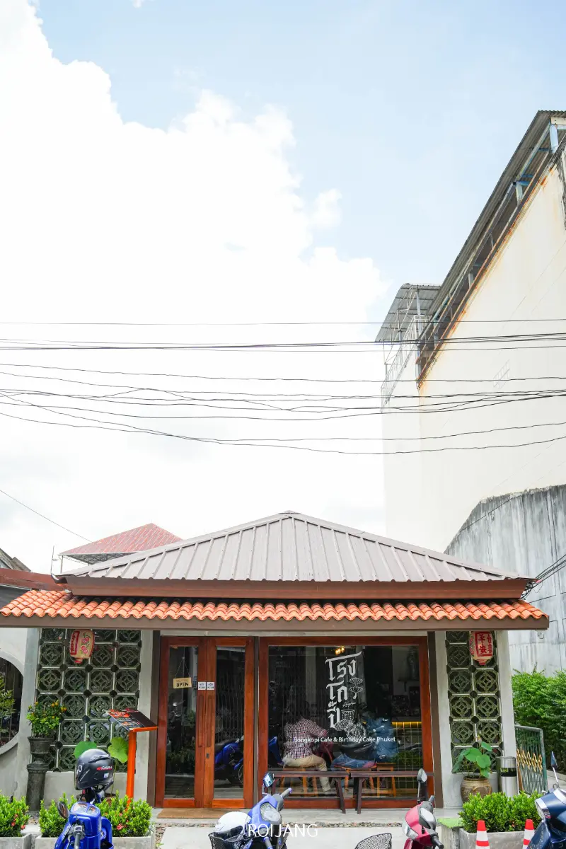 ร้านกาแฟเล็กๆ ชื่อโรงโกปี้ มีหลังคากระเบื้องสีน้ำตาลและด้านหน้าเป็นกระจก ตั้งอยู่ระหว่างอาคารขนาดใหญ่ 2 หลัง มีรถมอเตอร์ไซค์จอดอยู่ข้างนอกบนถนน.