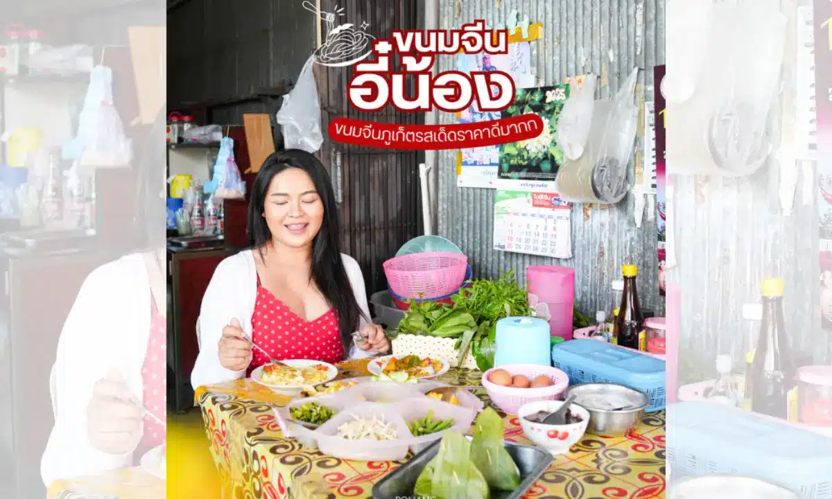 ผู้หญิงในชุดลายจุดสีแดงนั่งอยู่ที่โต๊ะที่มีอาหารไทยต่างๆ รวมทั้งขนมจีนอี๋น้อง ในบรรยากาศห้องครัวสบายๆ พร้อมปฏิทินและเครื่องปรุงเป็นพื้นหลัง.