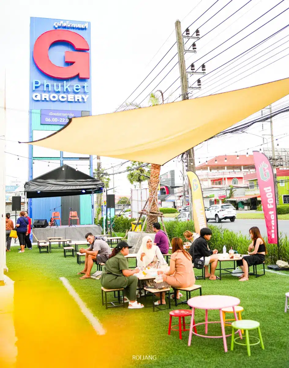 ผู้คนนั่งที่โต๊ะกลางแจ้ง กินอาหารและพูดคุยกันบนพื้นที่หญ้าใกล้กับร้านขายของชำโกรเซอรี่ภูเก็ต โดยมีธงสีสันสดใสและป้ายร้านค้าขนาดใหญ่ที่มองเห็นได้ในพื้นหลัง.