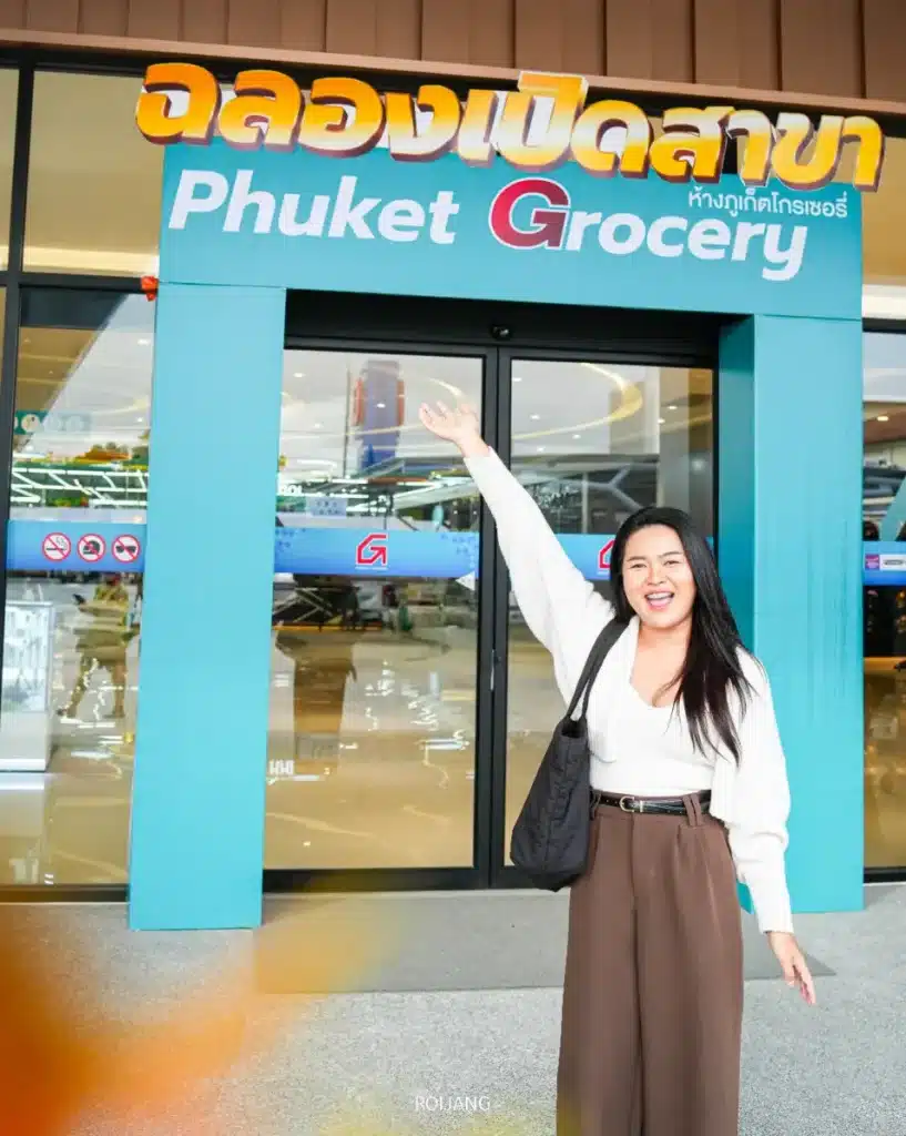ผู้หญิงคนหนึ่งยืนยิ้มพร้อมยกแขนข้างหนึ่งขึ้นหน้าทางเข้าร้านชื่อ "ภูเก็ต โกรเซอรี่.