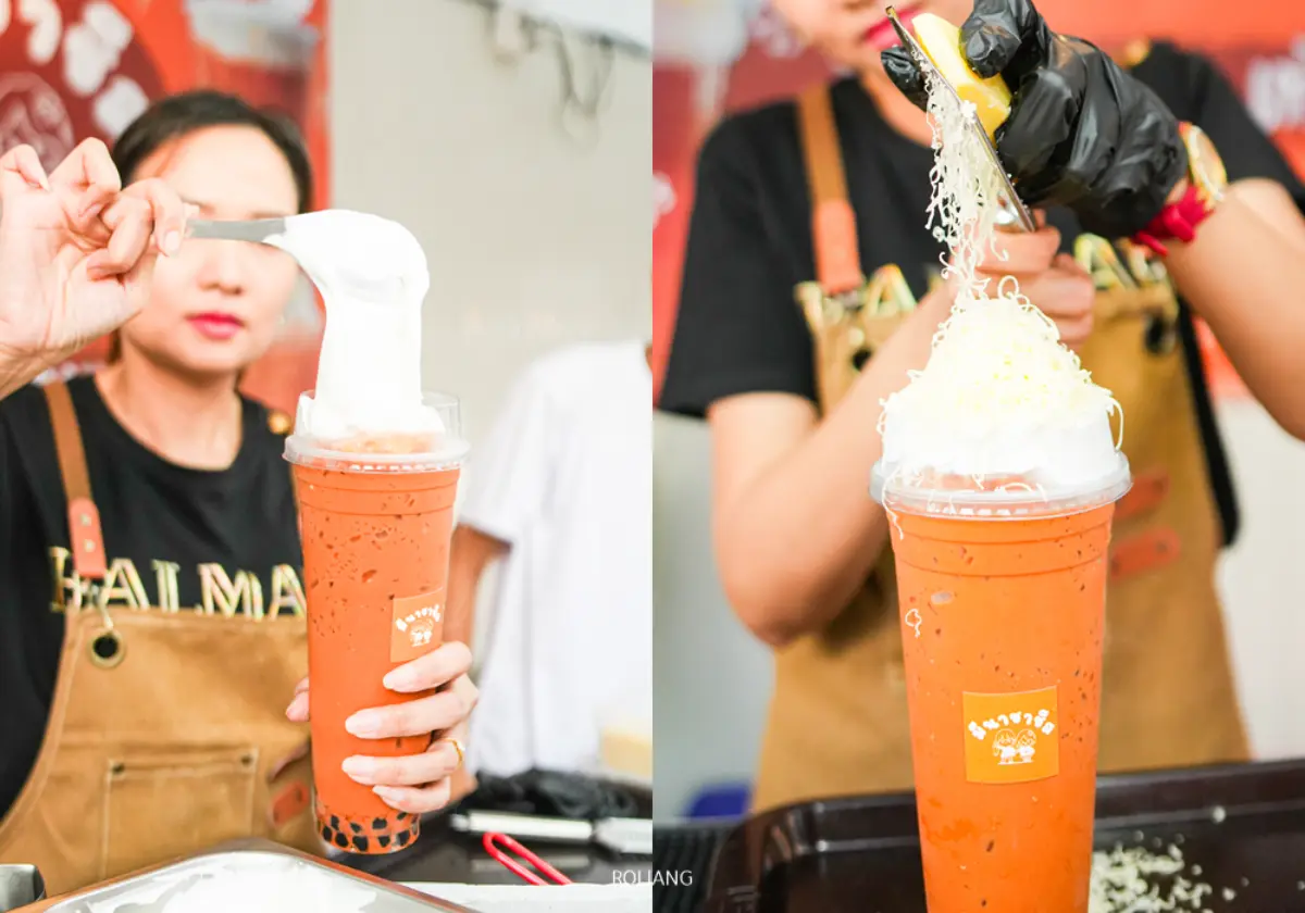 ชายคนหนึ่งกำลังเตรียมเครื่องดื่มชาเย็นแก้วใหญ่ราดด้วยวิปครีมและชีสขูดในร้านโกรเซอรี่สุดอบอุ่นแห่งหนึ่ง โดยเขาสวมผ้ากันเปื้อนและถุงมือ.