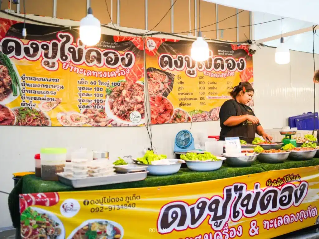 แผงขายอาหารที่มีคนสองคนเตรียมอาหาร วัตถุดิบต่างๆ วางอยู่บนเคาน์เตอร์ และมีแบนเนอร์ภาษาไทยสีสันสวยงามของห้างโกรเซอรี่ที่จัดแสดงอยู่เบื้องหลัง.