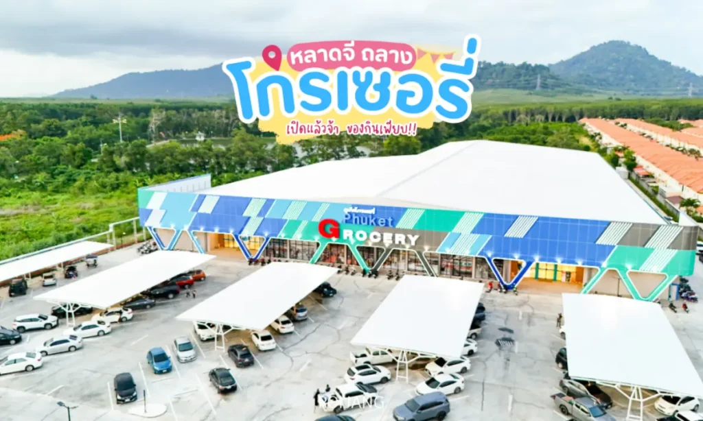 มุมมองทางอากาศของตลาดภูเก็ตโกรเซอรี่ที่มีลานจอดรถขนาดใหญ่ที่เต็มไปด้วยรถยนต์ ภายนอกที่ทันสมัย และมีฉากหลังเป็นภูเขา.
