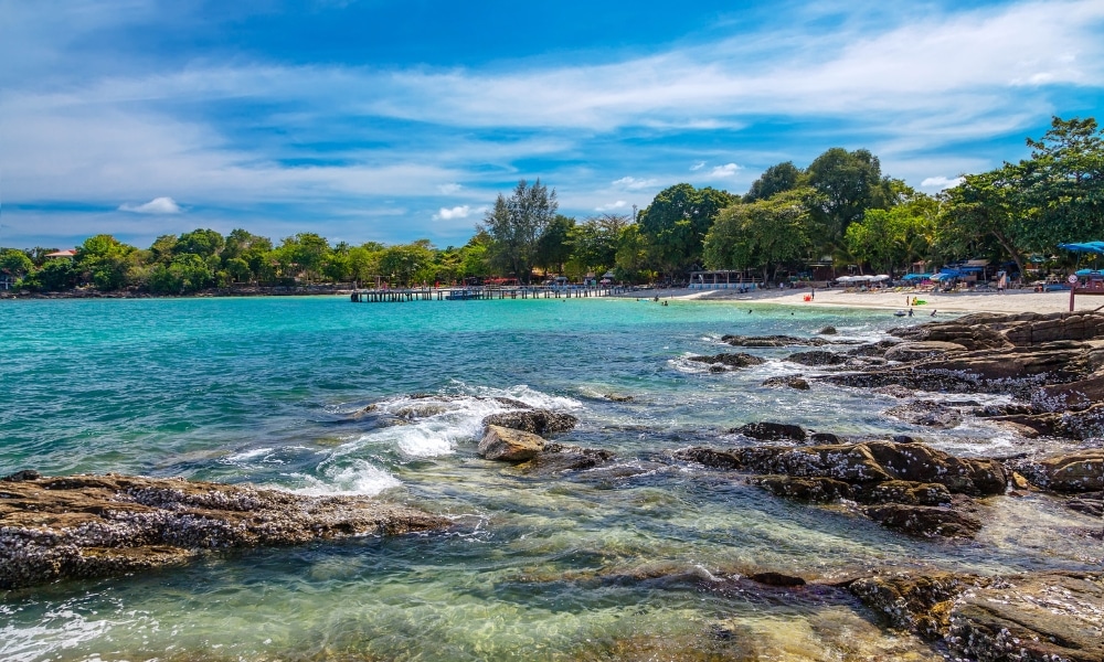น้ำทะเลสีฟ้าใสไหลผ่านชายฝั่งหิน มีชายหาดทรายและต้นไม้สีเขียวเป็นฉากหลังภายใต้ท้องฟ้าสีฟ้าที่มีเมฆบางส่วน เหมาะสำหรับผู้ที่ต้องการ ที่เที่ยวเกาะเสม็ด สำรวจ ที่เที่ยวเกาะเสม็ด ในการผจญภัยครั้งต่อไป.