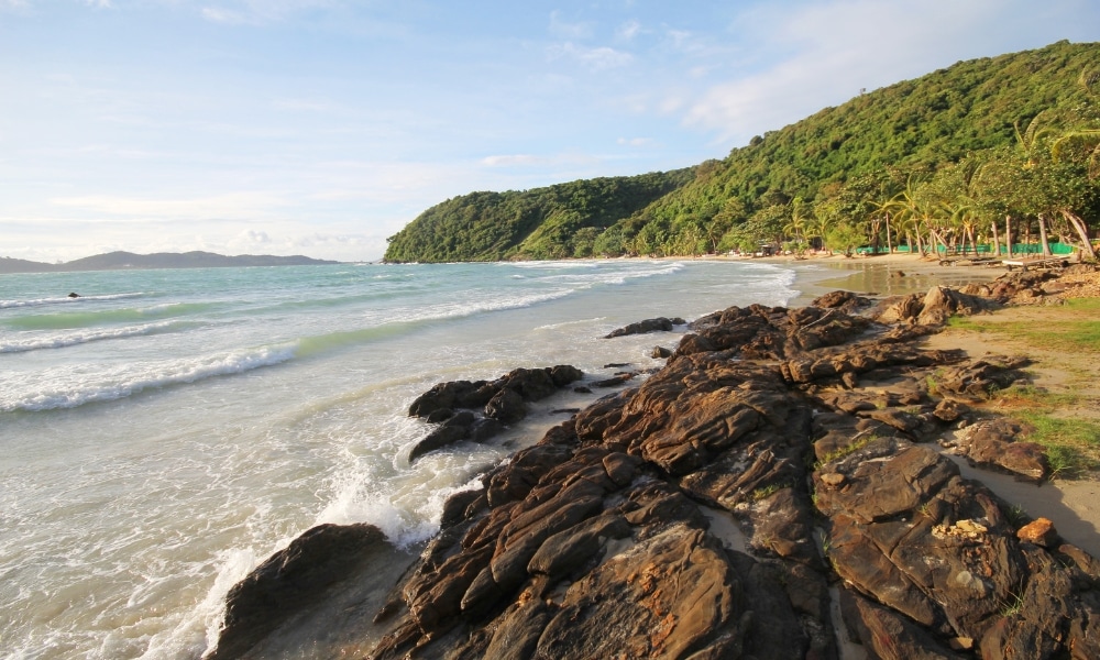ชายฝั่งหินที่มีคลื่นอ่อนโยน หาดทราย ต้นปาล์ม และเนินเขาเขียวขจีภายใต้ท้องฟ้าที่มีเมฆบางส่วน ที่เที่ยวเกาะเสม็ด  เหมาะสำหรับผู้ที่มองหาเที่ยวเกาะเสม็ดและค้นพบความงามทางธรรมชาติของที่เที่ยวเกาะเสม็ด.