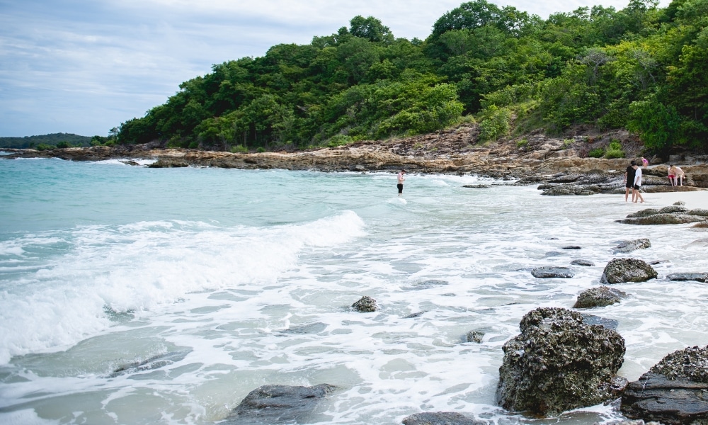 หาดทรายที่มีหินกระจัดกระจาย คลื่นเบาๆ และผู้คนริมชายฝั่ง มีต้นไม้เขียวขจีปกคลุมเนินเขาเป็นฉากหลัง เหมาะสำหรับผู้ที่มองหา ที่เที่ยวเกาะเสม็ด