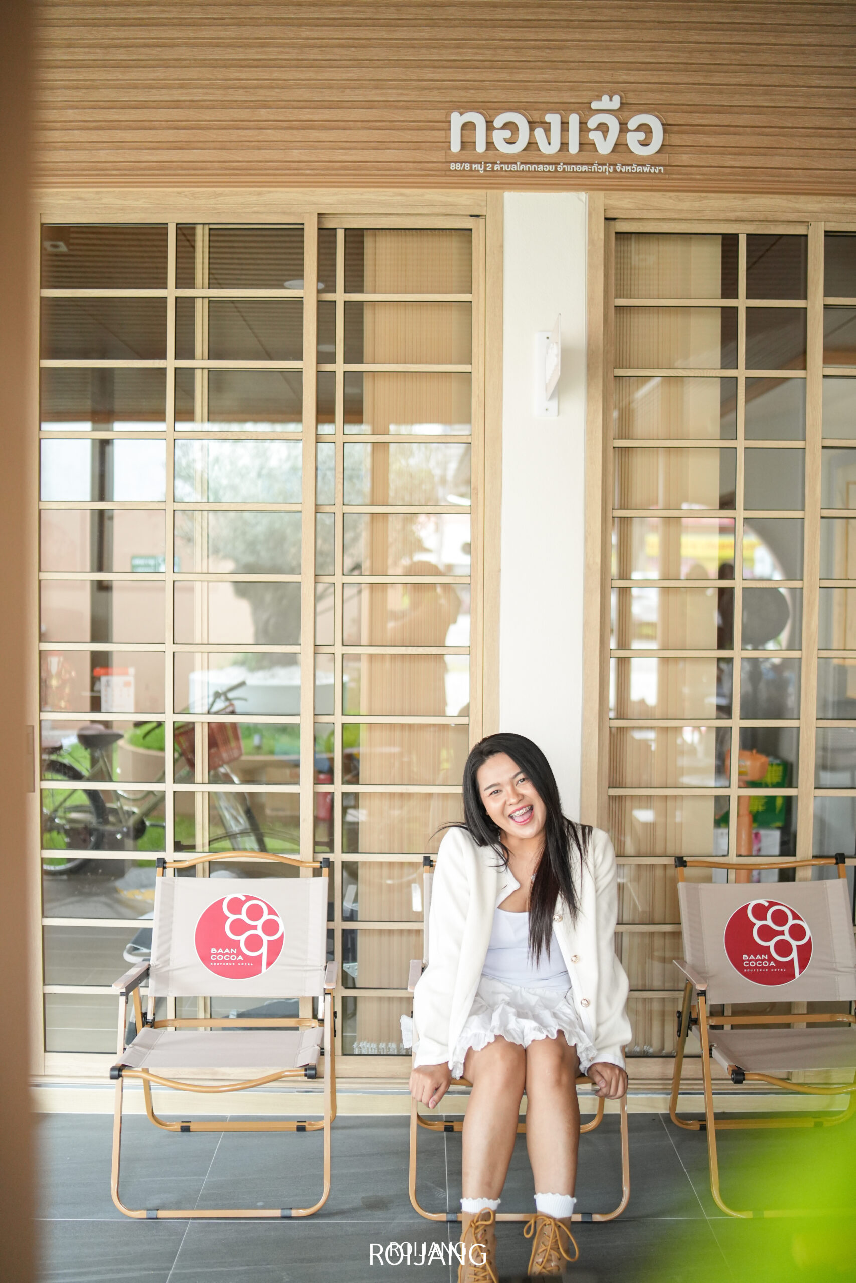 ผู้หญิงในชุดสีขาวนั่งและยิ้มอยู่บนเก้าอี้หน้าบ้านโกโก้ ซึ่งเป็นอาคารที่มีป้ายภาษาไทย โดยมีเก้าอี้ว่างเปล่าสองตัวอยู่ด้านข้าง.