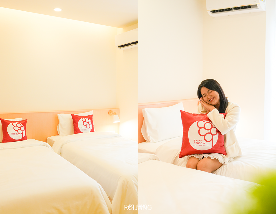 ห้องพักในโรงแรมบ้านโกโก้ มีเตียงเดี่ยว 2 เตียงและชุดเครื่องนอนสีขาวสะอาด มีผู้หญิงนั่งอยู่บนเตียงหนึ่งเตียง พร้อมถือหมอนสีแดงที่มีโลโก้และข้อความ.
