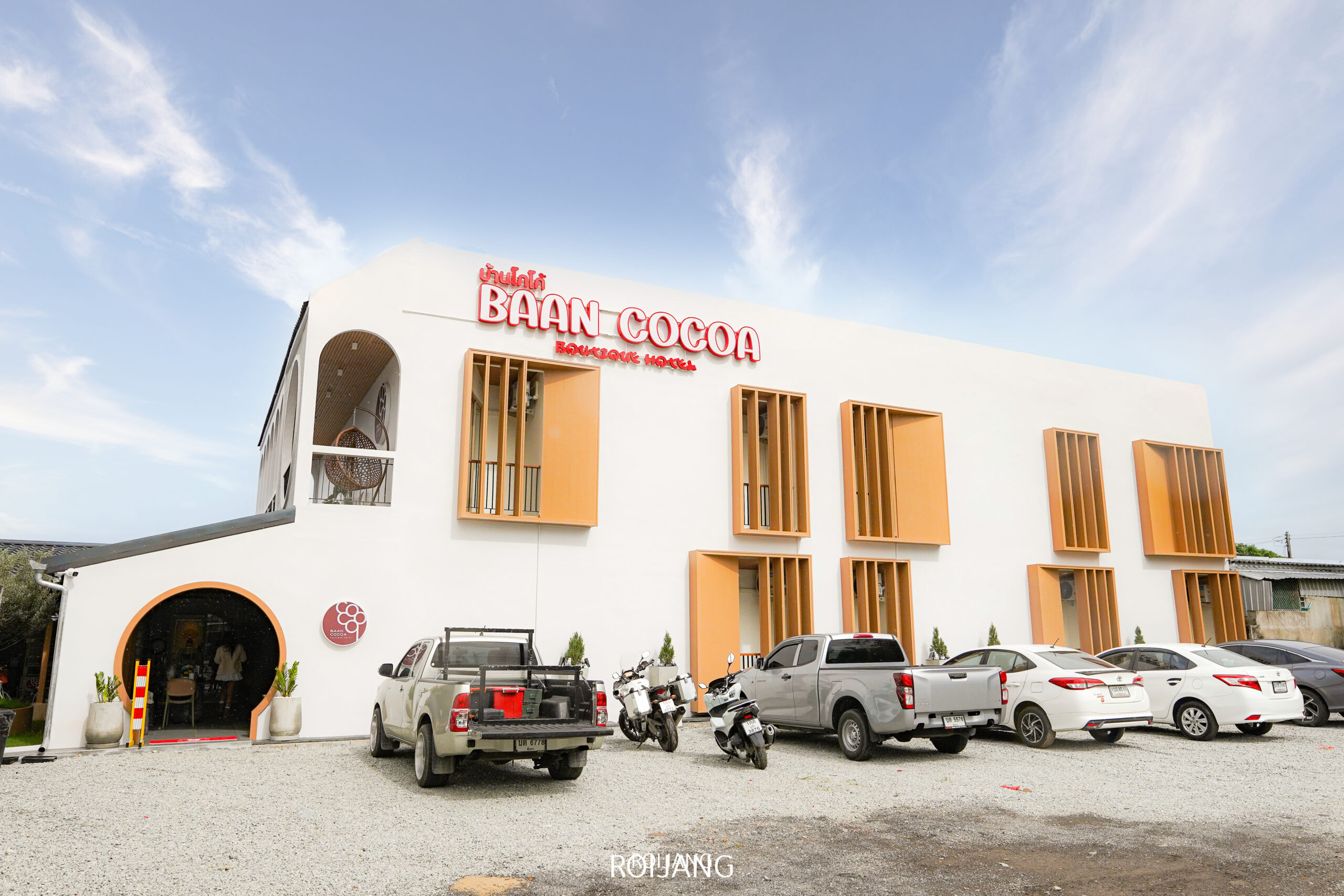 อาคารสีขาว 2 ชั้นสไตล์โมเดิร์นที่มีป้ายว่า “Baan Cocoa Boutique Hotel” ตั้งอยู่ท่ามกลางท้องฟ้าแจ่มใส พร้อมด้วยรถยนต์และรถจักรยานยนต์หลายคันจอดอยู่ด้านหน้า คอยต้อนรับแขกผู้มาเยือนสู่ประสบการณ์ Baan Cocoa สุดเก๋.