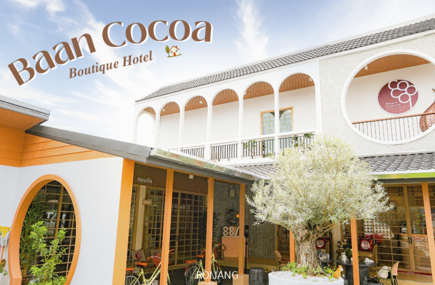 ด้านหน้าของโรงแรม Baan Cocoa Boutique Hotel โดดเด่นด้วยซุ้มประตูสีขาว ตกแต่งด้วยสีส้ม ต้นไม้ใหญ่ และป้าย Baan Cocoa ที่ตั้งโดดเด่นอยู่เหนืออาคาร.