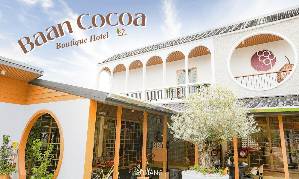 ด้านหน้าของโรงแรม Baan Cocoa Boutique Hotel โดดเด่นด้วยซุ้มประตูสีขาว ตกแต่งด้วยสีส้ม ต้นไม้ใหญ่ และป้าย Baan Cocoa ที่ตั้งโดดเด่นอยู่เหนืออาคาร.