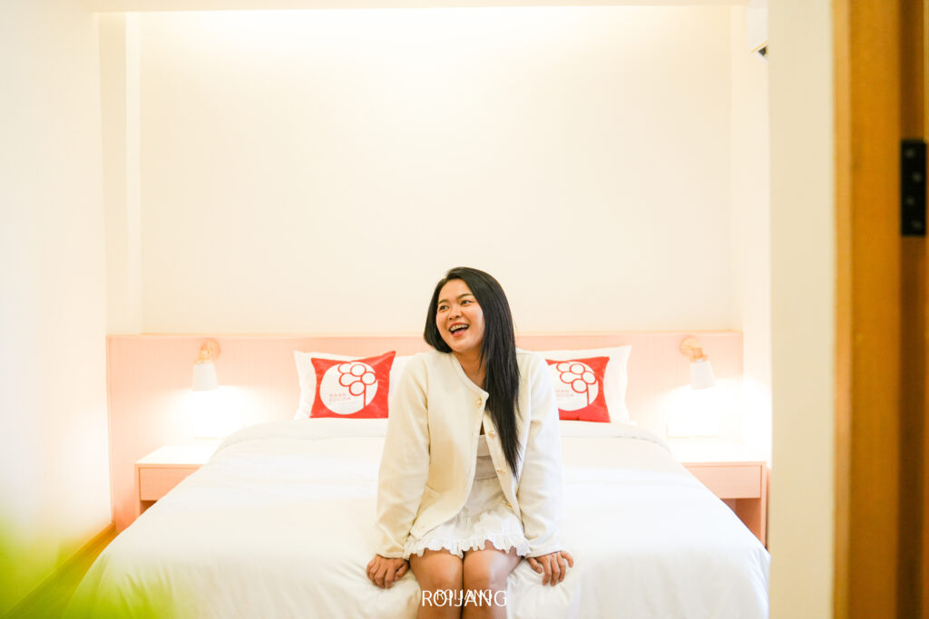 ผู้หญิงคนหนึ่งนั่งอยู่บนเตียงที่จัดอย่างเรียบร้อยในห้องนอนบ้านโกโก้ที่สว่างสดใสและเรียบง่าย เธอยิ้มและมองไปด้านข้าง.