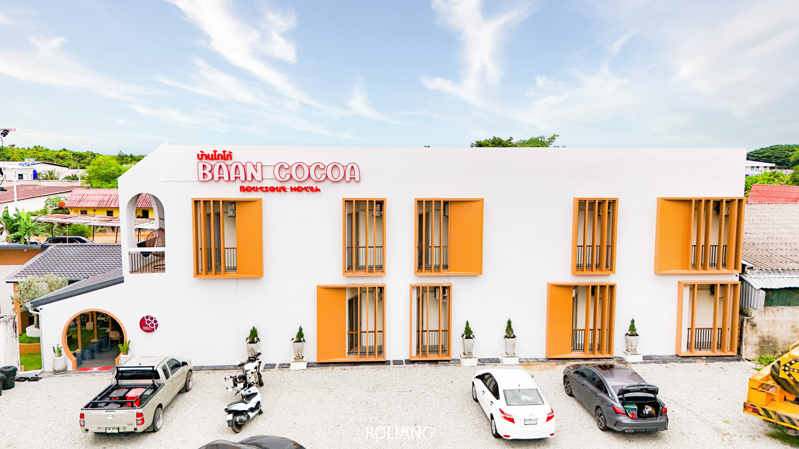 อาคารสีขาวสไตล์โมเดิร์นที่มีป้ายว่า “Baan Cocoa Boutique Hotel” พร้อมด้วยหน้าต่างสีส้ม รถจอดอยู่หลายคัน และท้องฟ้าที่มีเมฆบางส่วน สะท้อนถึงเสน่ห์อันน่าดึงดูดของ Baan Cocoa.