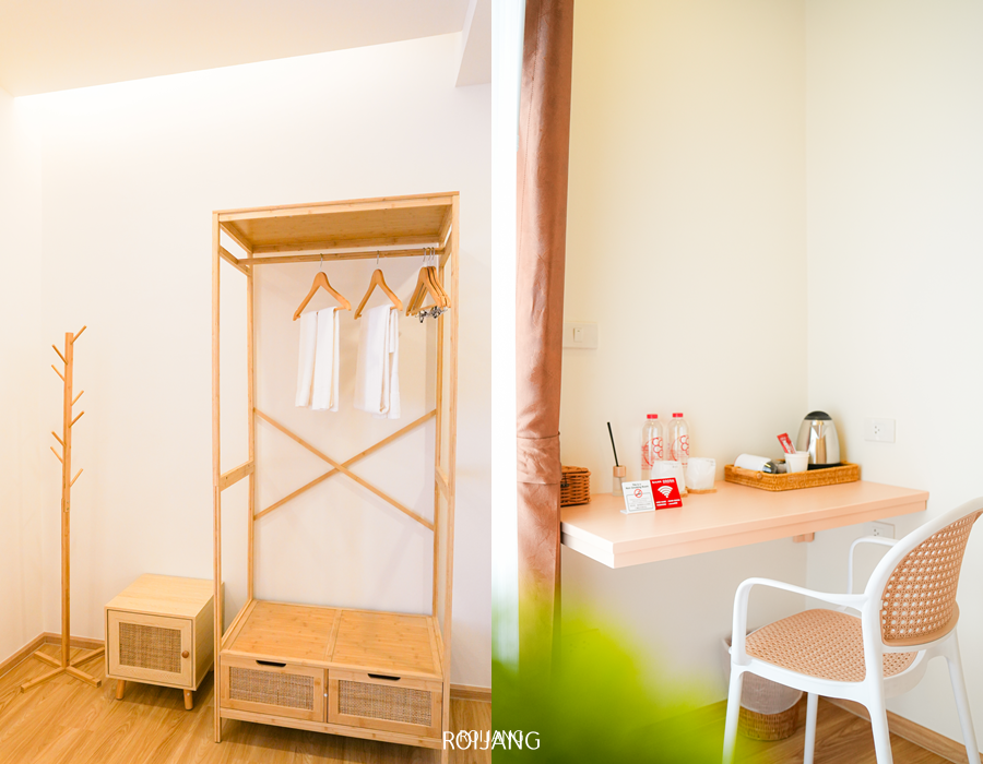 มุมห้องพักโรงแรมบ้านโกโก้สไตล์มินิมอล มีตู้เสื้อผ้าไม้แบบเปิดและราวแขวนเสื้ออยู่ทางซ้าย โต๊ะทำงานที่มีเครื่องดื่ม กาต้มน้ำ และเก้าอี้อยู่ทางขวา.