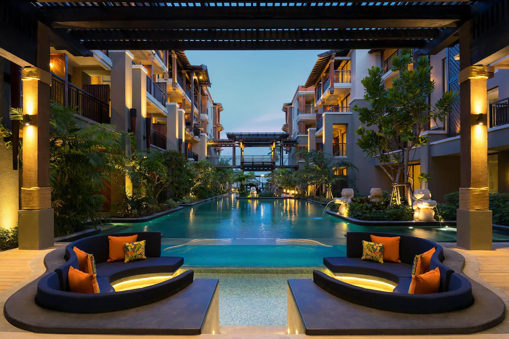 บริเวณสระว่ายน้ำของโรงแรมทันสมัยในยามพลบค่ำพร้อมเก้าอี้เลานจ์ที่จมลง หมอนอิงตกแต่ง ต้นไม้เมืองร้อน และระเบียงหลายชั้นที่มองเห็นสระว่ายน้ำที่มีแสงไฟส่องสว่าง เหมาะสำหรับการผ่อนคลายหลังจากใช้เวลาทั้งวันใน การท่องเที่ยวเกาะสมุย.