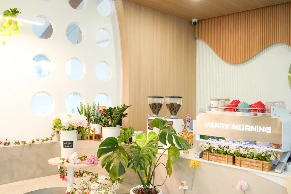ร้าน Merry Morning Cafe มีการตกแต่งภายในที่สดใสและทันสมัย พร้อมด้วยต้นไม้เขียวชอุ่ม ดอกไม้สด เครื่องบดกาแฟ 2 เครื่อง และเครื่องชงเอสเพรสโซที่มีป้ายติดว่า "Merry Morning.