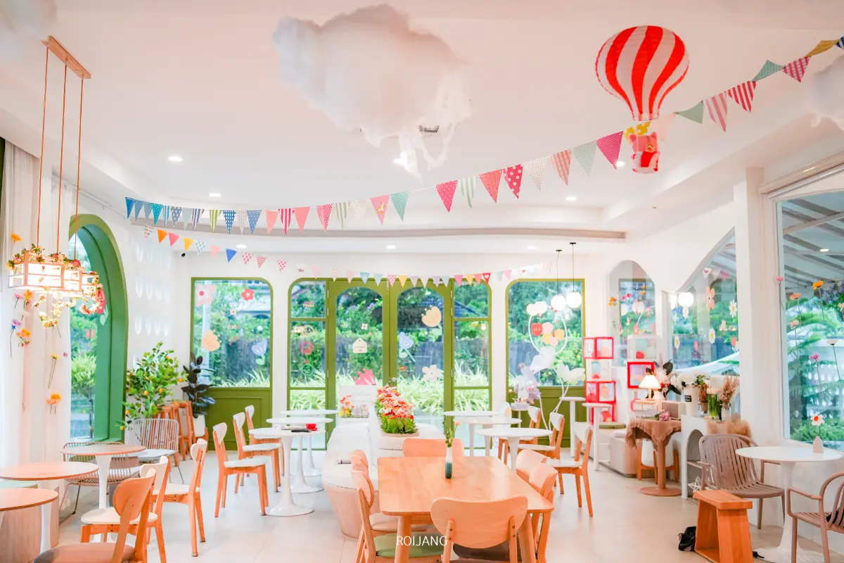 ภายในคาเฟ่ Merry Morning Cafe ที่มีสีสันสดใส ตกแต่งด้วยธงหลากสี ลูกโป่ง และก้อนเมฆ โต๊ะและเก้าอี้ไม้เต็มไปหมด ขณะที่หน้าต่างบานใหญ่เผยให้เห็นต้นไม้เขียวชอุ่มด้านนอก.
