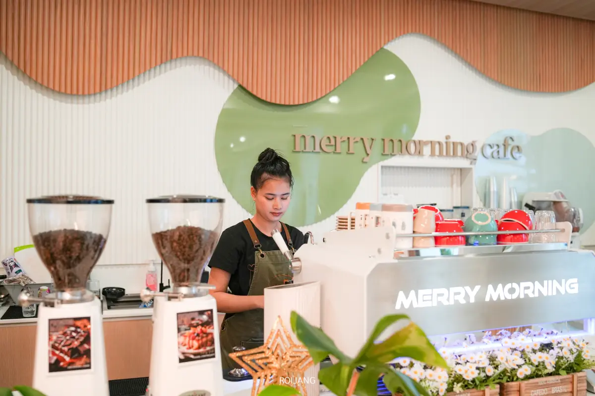 บาริสต้าของร้าน Merry Morning Cafe กำลังเตรียมกาแฟอยู่หลังเคาน์เตอร์ ซึ่งรายล้อมไปด้วยเครื่องชงกาแฟ ถ้วยกาแฟ และเมล็ดกาแฟ ในบรรยากาศที่ทันสมัยและมีแสงสว่างเพียงพอ.