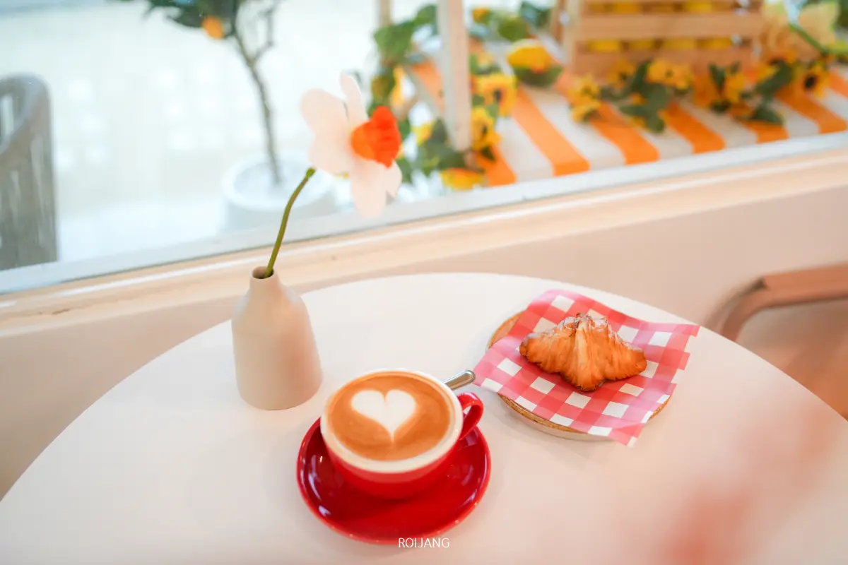 ที่ร้าน Merry Morning Cafe มีแก้วลาเต้ที่มีลายลาเต้รูปหัวใจในแก้วสีแดงวางอยู่ข้างๆ ครัวซองต์บนผ้าเช็ดปากลายตารางหมากรุก และมีดอกไม้สีขาวในแจกันช่วยเติมเต็มบรรยากาศอบอุ่นนี้ให้สมบูรณ์แบบ.