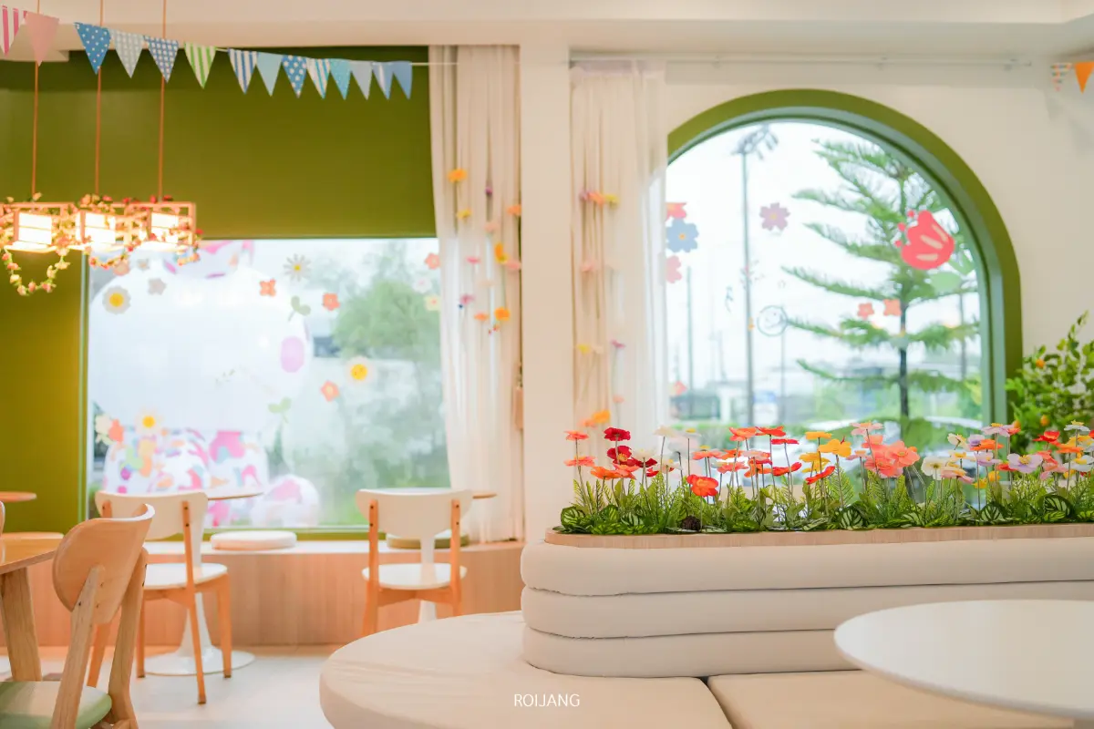 บรรยากาศภายในร้าน Merry Morning Cafe ที่สดใสและอบอุ่น มีทั้งเก้าอี้ไม้ โต๊ะกลม หน้าต่างกรอบสีเขียวขนาดใหญ่ และดอกไม้ประดิษฐ์หลากสีสันเป็นของตกแต่ง มีธงและดอกไม้ประดับห้อยลงมาจากเพดาน.