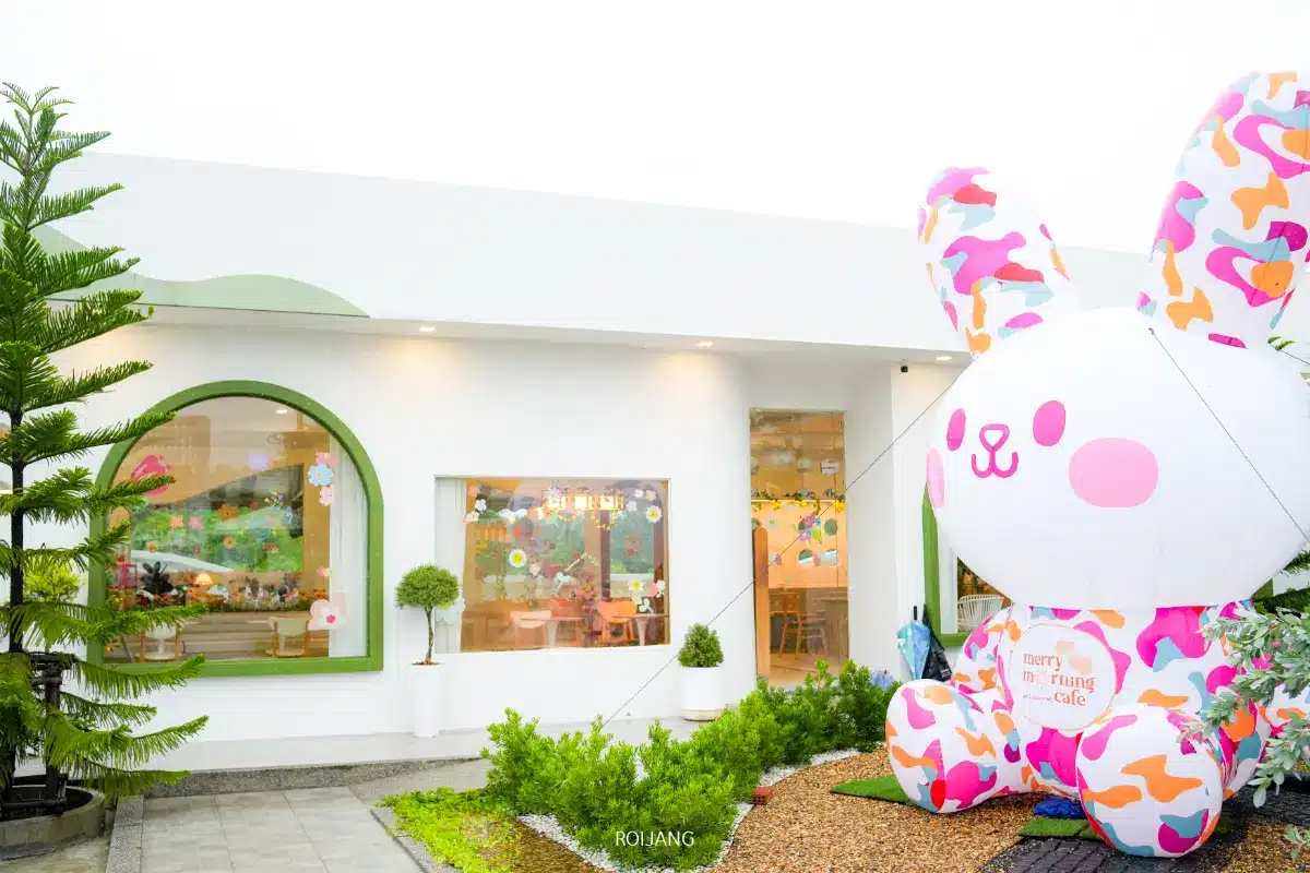 อาคารสีขาวที่มีหน้าต่างบานใหญ่และขอบสีเขียวซึ่งเป็นที่ตั้งของ Merry Morning Cafe ตั้งอยู่ติดกับกระต่ายเป่าลมสีสันสดใสขนาดยักษ์ที่ทางเข้า ต้นไม้และพุ่มไม้ช่วยเพิ่มเสน่ห์ให้กับฉากที่มีชีวิตชีวา.